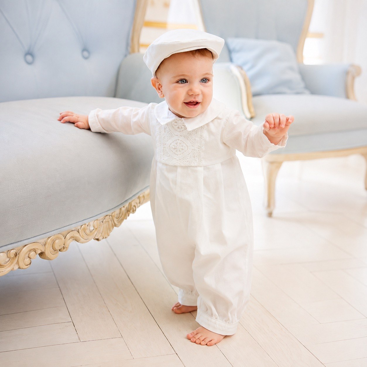 A baby wearing a white long-sleeve baby romper with lace embroidery on the chest and a matching white baby hat, standing on a light wood floor in a bright room.