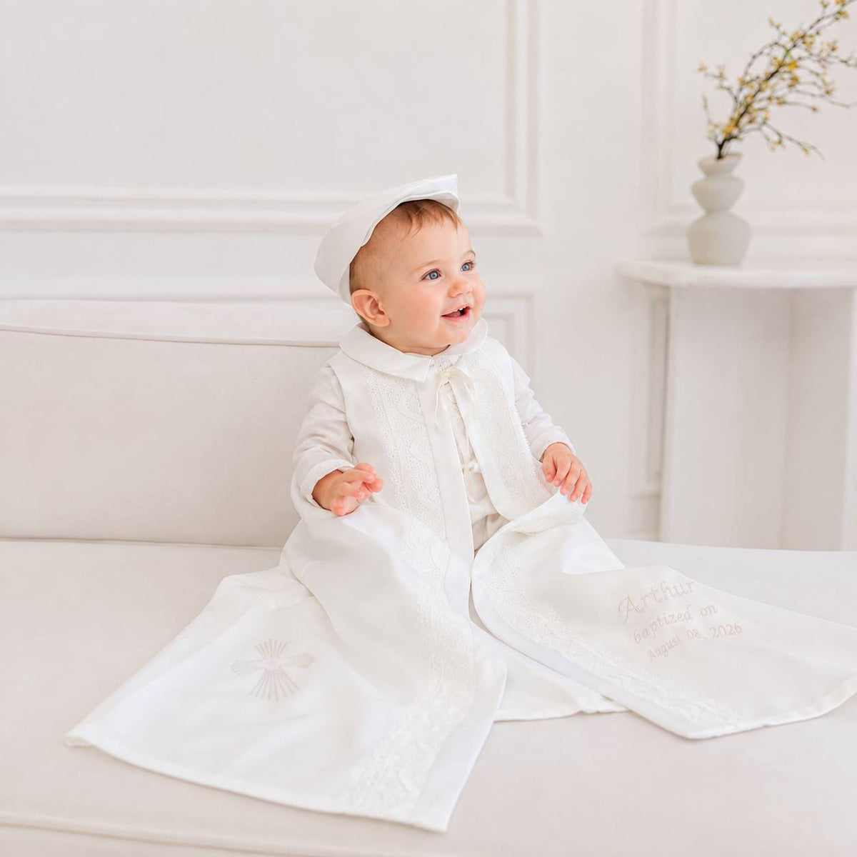 A baby sitting on a white sofa wearing a white long-sleeve christening gown with a matching bonnet. The gown has a high collar, long sleeves, and embroidered details on the front and sleeves.