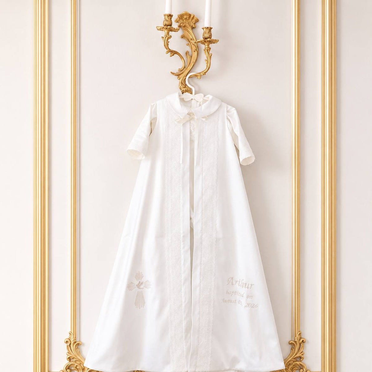 A white baby christening gown with short sleeves, a ruffled collar, and delicate gold embroidery, hanging from a gold wall-mounted candlestick on a cream-colored paneled wall.