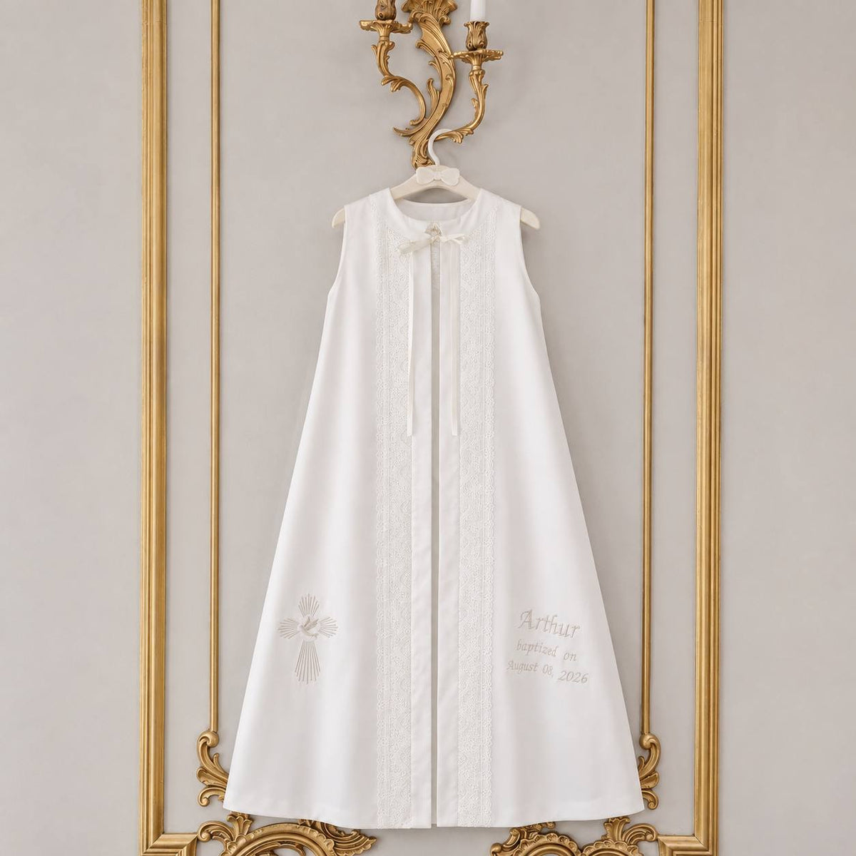 A sleeveless baptism robe with lace trim and a small bow at the neckline, hanging on a hanger against a decorative gold-framed wall.