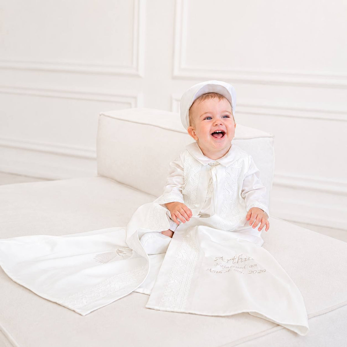 A smiling baby wearing a white first baptism robe with a matching bonnet sits on a white upholstered bench. The outfit includes a long-sleeve tunic with a bow tie and a long embroidered robe with the name 'Arthur' and 'First Communion 2020' embroidered on the front.