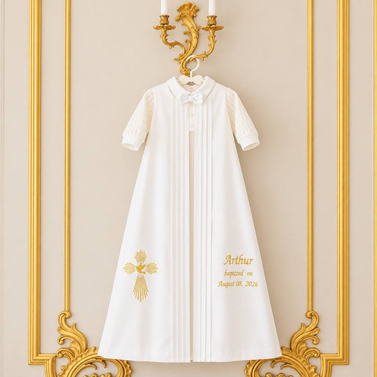 A white short-sleeve baby baptism gown with gold embroidery, hanging on a hanger against an ornate gold-trimmed beige wall. The gown features a pleated front, a white collar with a bow, and custom gold text reading 'Arthur baptized on August 08, 2026' on the lower right side.
