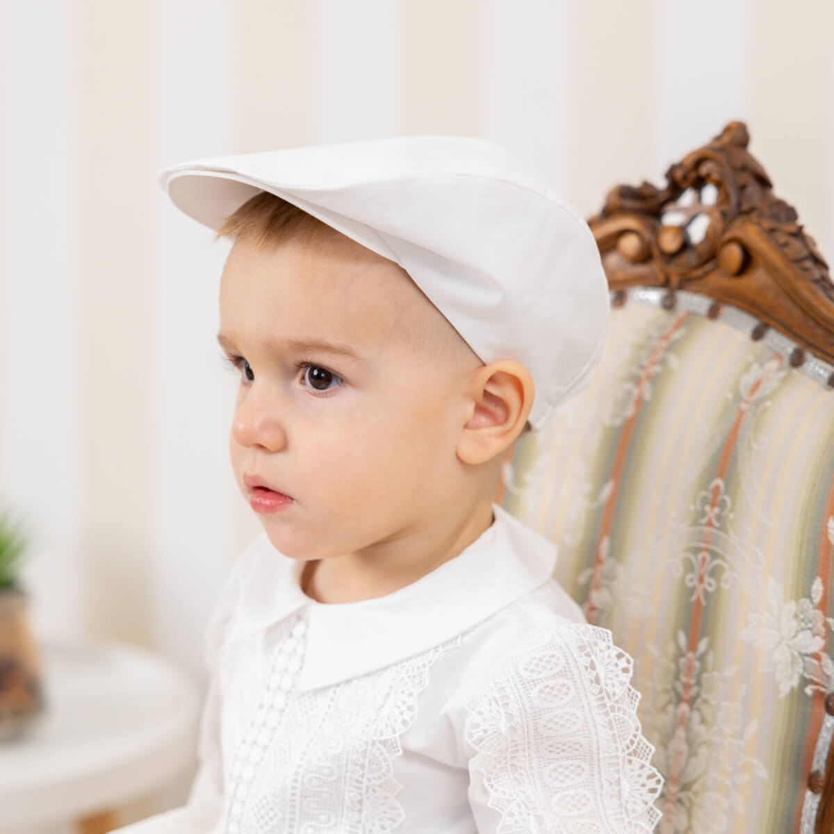 Child wearing a white outfit with a matching hat in a softly blurred indoor setting
