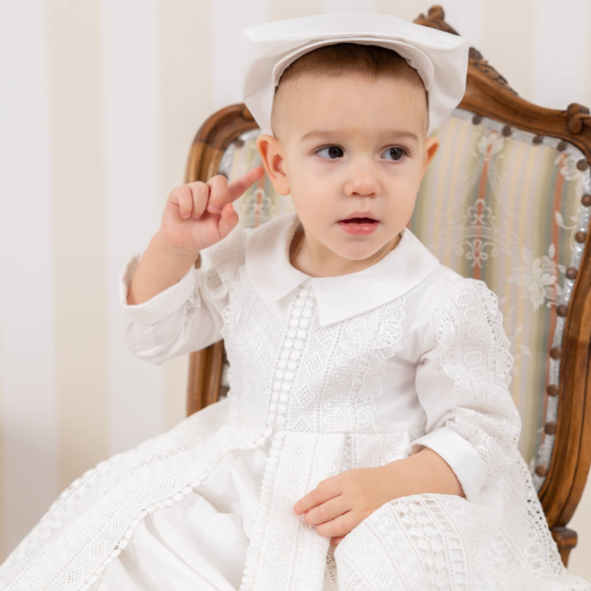 Child in a christening outfit with a hat sitting on a decorative chair.