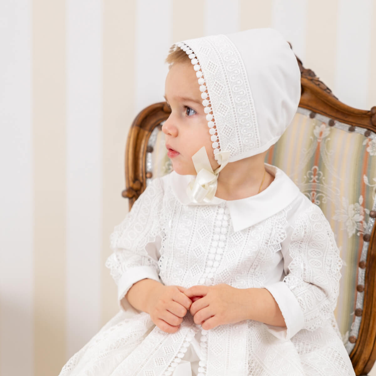 Boy wearing a christening gown and bonnet sitting on a wooden chair.