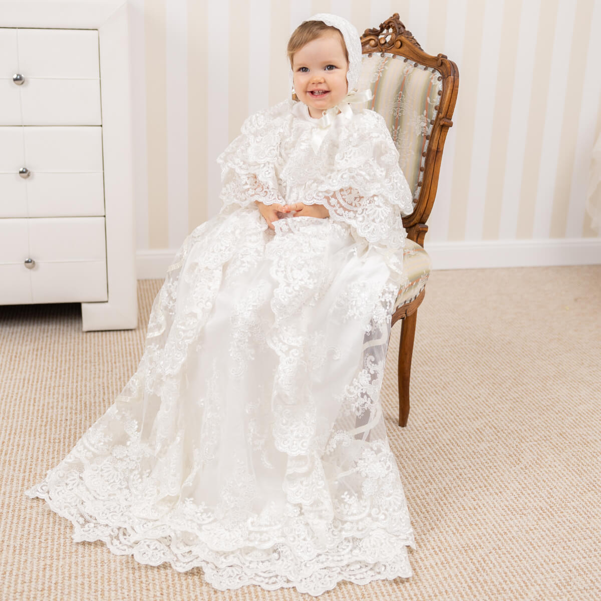Baby in a white lace christening gown sitting on a wooden chair in a room 