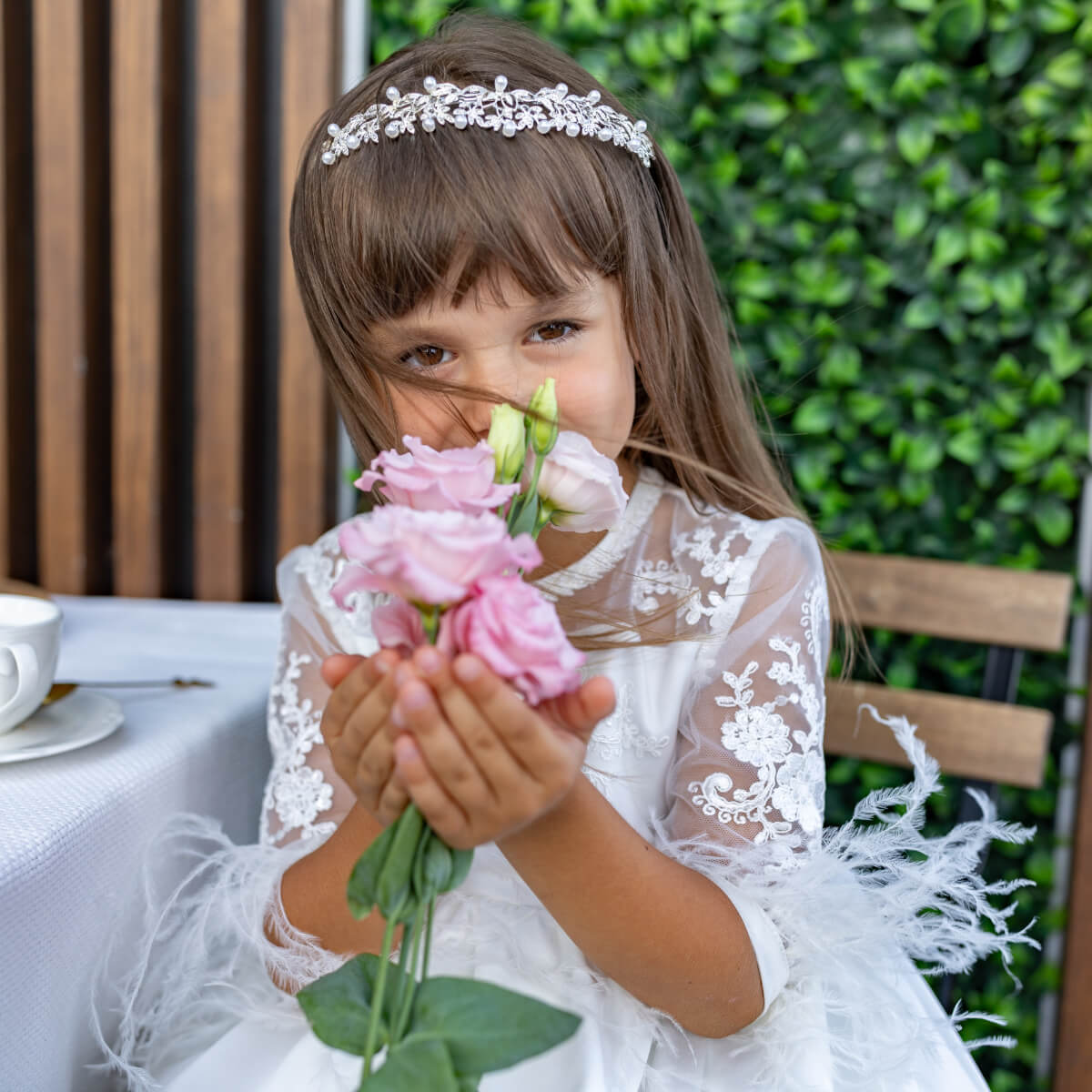 Young girl in a white lace dress holding flowers outdoors with greenery in the background