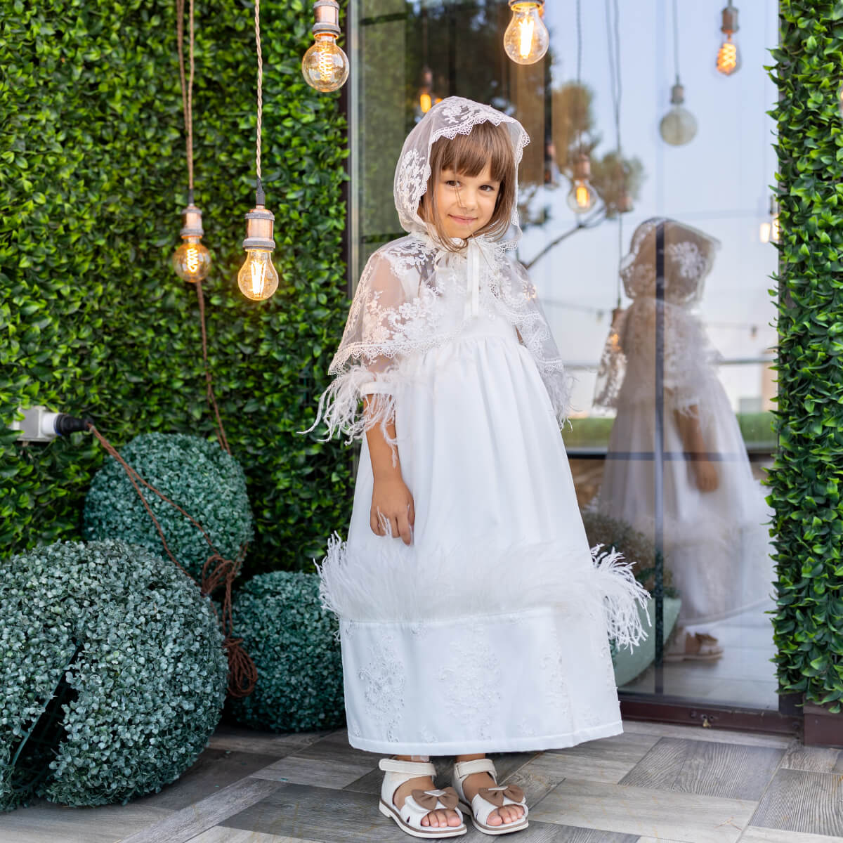 Young girl in a Luxury Communion Dress with a hood standing in front of a store window with greenery.