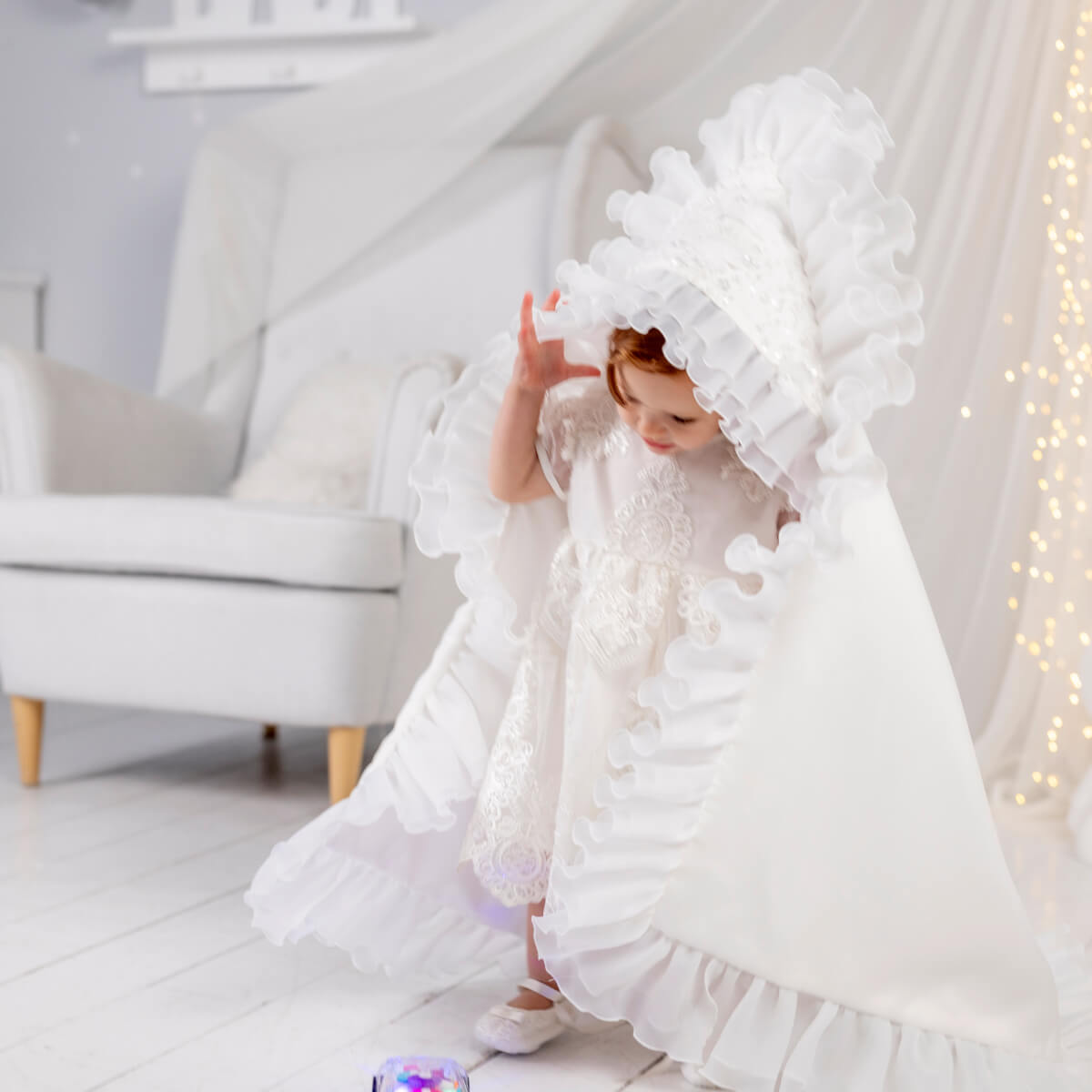 Child in a whit luxury blanket for baptism standing in a bright room with a white chair and decorative lights.