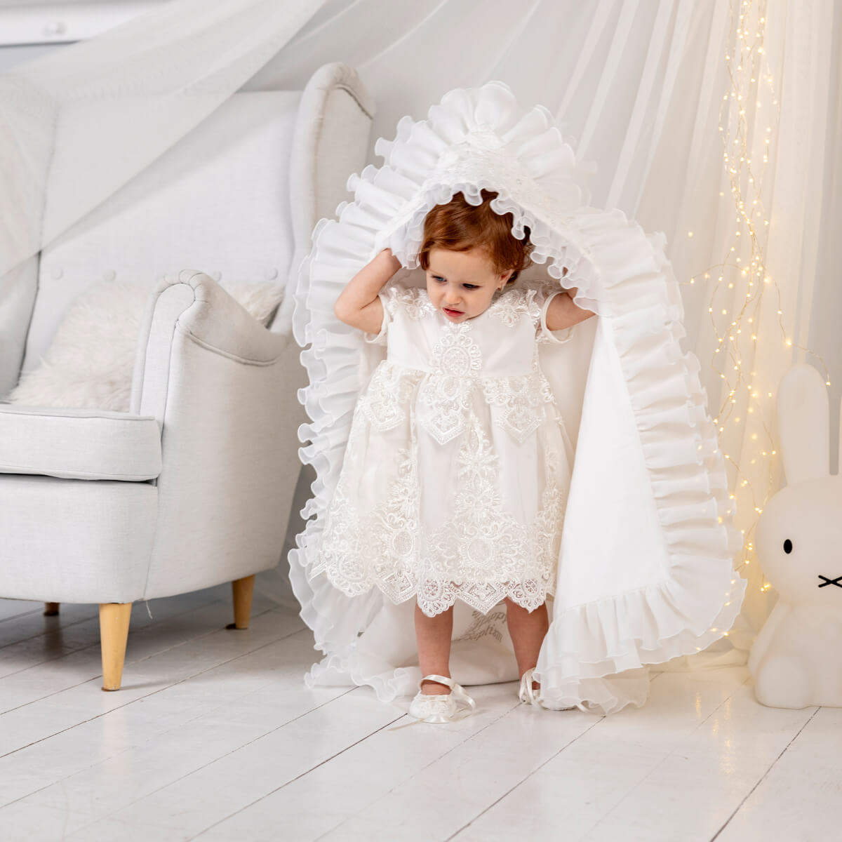 Child in a white  luxury blanket for baptism standing under a decorative arch in a bright room.