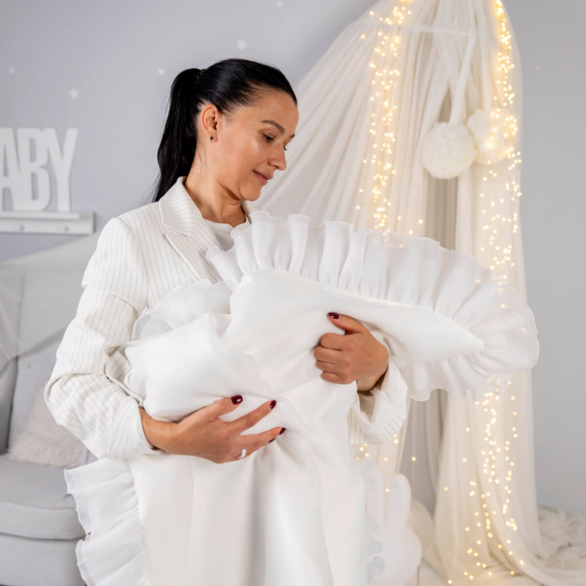 Woman holding a baby wrapped in a white blanket with decorative lights in the background