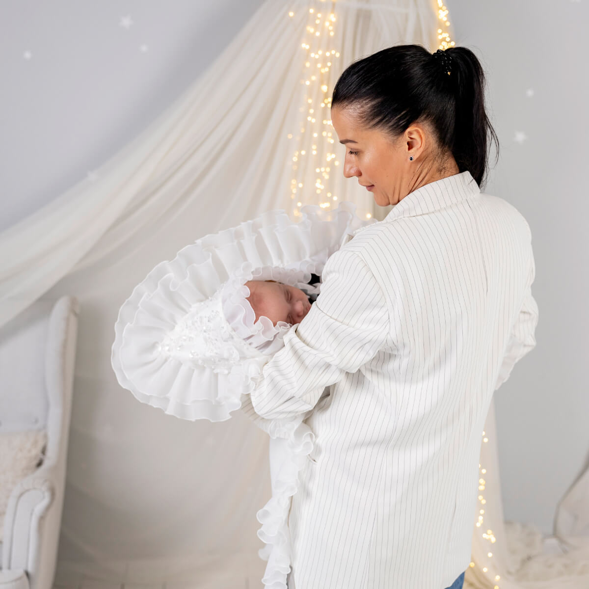 Woman holding a baby in a crib with white bedding and fairy lights in the background