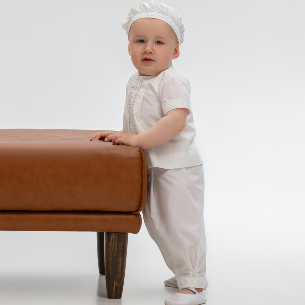 Baby in Baby Boy Baptism Suit standing next to a brown leather ottoman on a white background