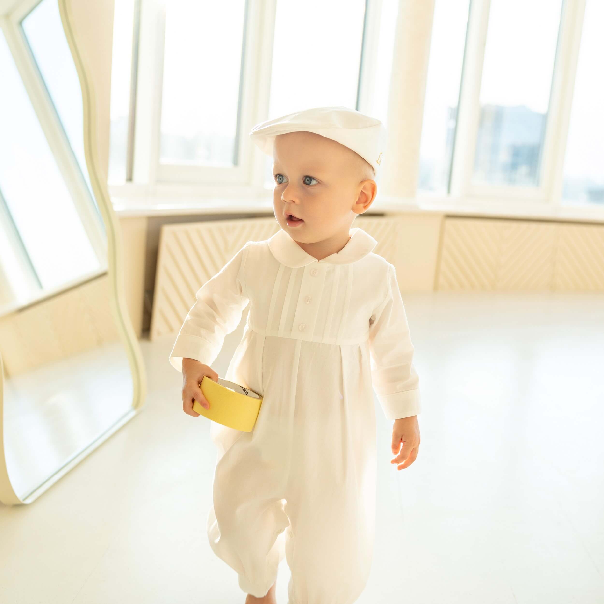 Baby in a Baby Boy Baptism Bubble Romper standing in a bright room with large windows.