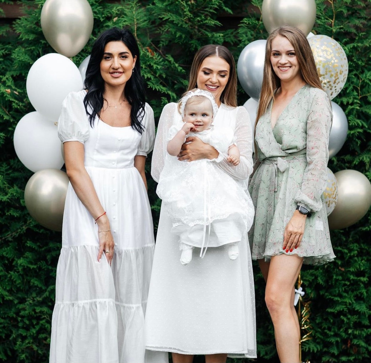 Three women in formal attire with a baby, wearing Lace Baptism Set for Baby Girl surrounded by balloons and greenery