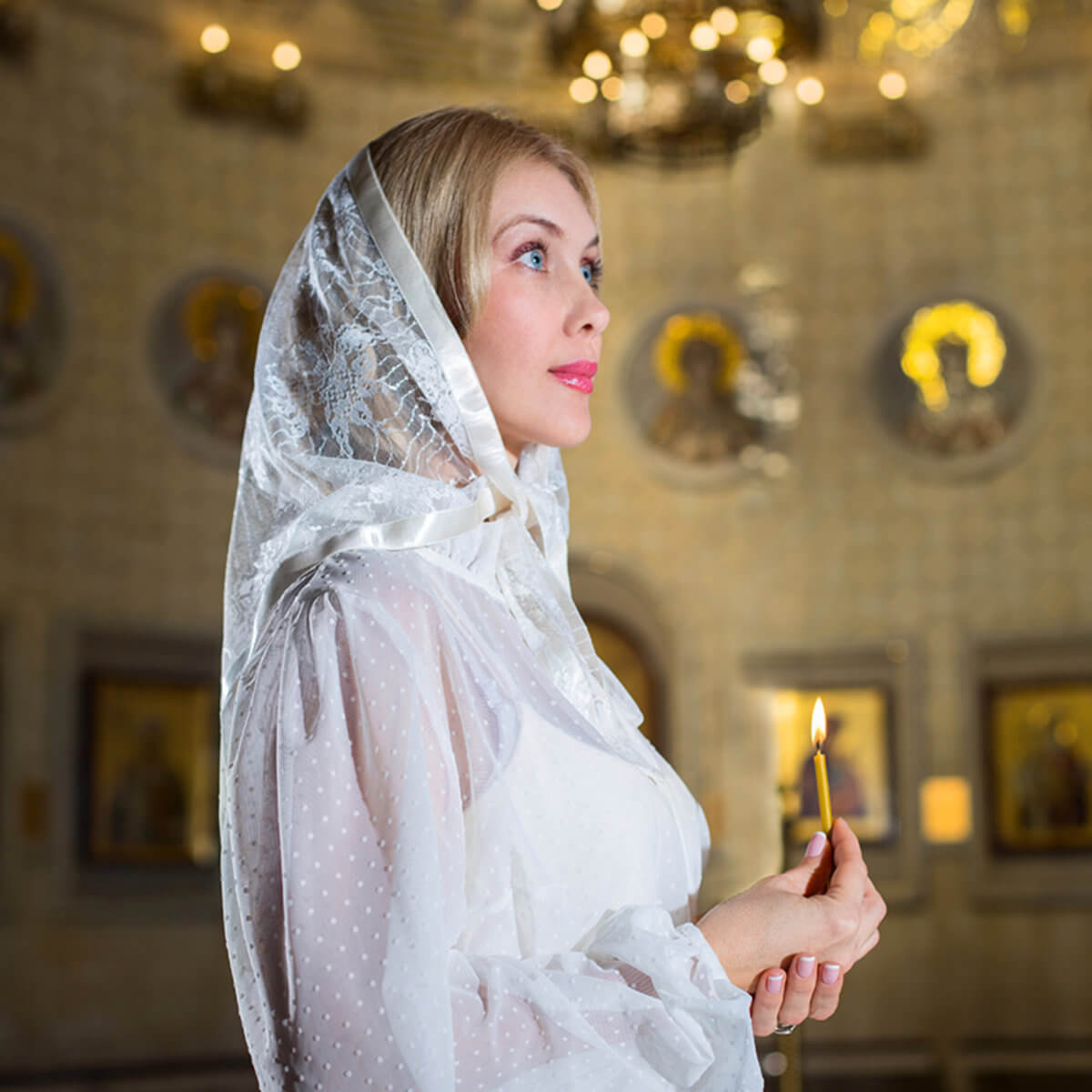 Woman in a white dress and in Lace Chapel Veil holding a lit candle in an ornate interior setting