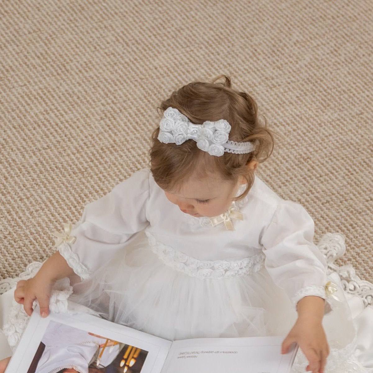Baby in a white dress baby headband with bow sitting on a textured surface.