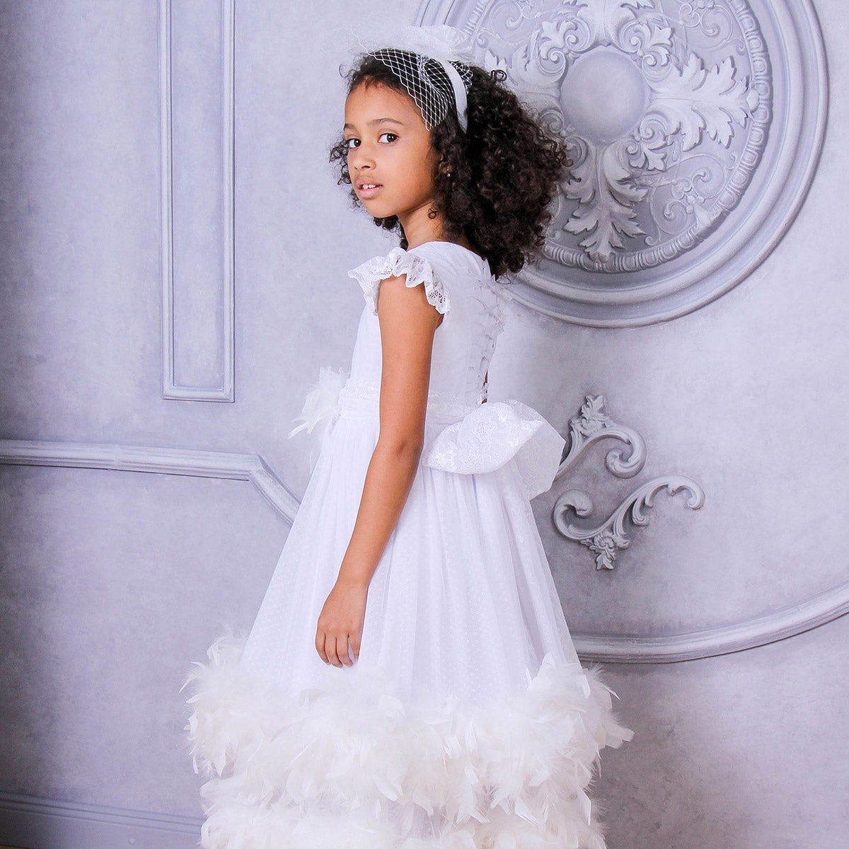 Young girl in a white dress standing against an ornate wall.