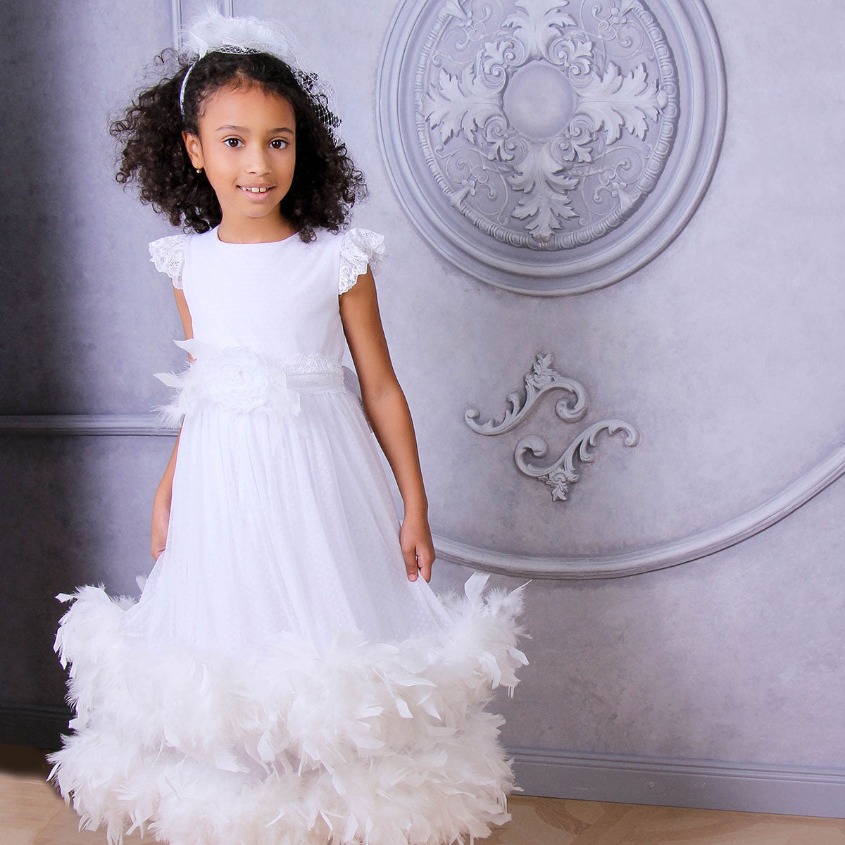 Young girl in a white dress with ruffled sleeves and feathered skirt standing against an ornate wall.