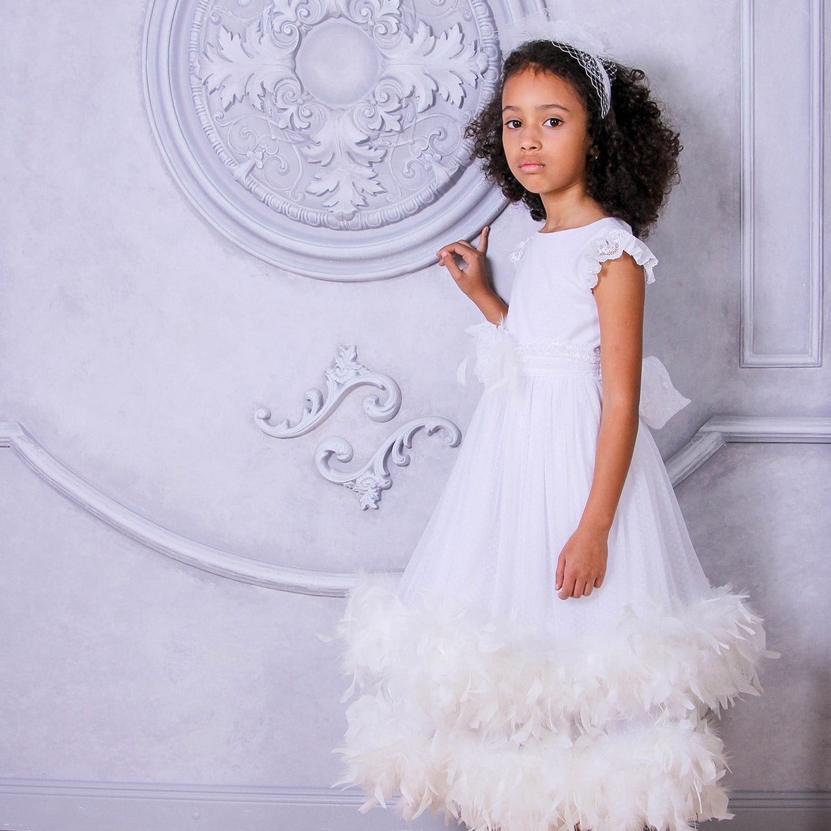 Young girl in a white dress with feather details against an ornate wall.
