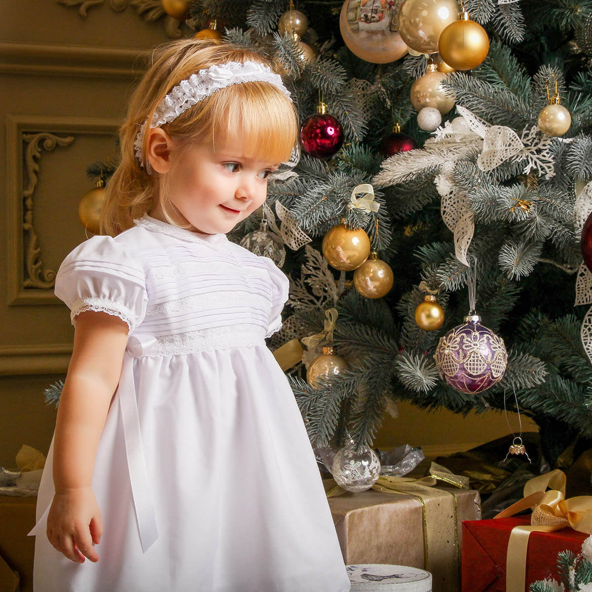 A child inCotton Christening Dress standing in front of a decorated Christmas tree with ornaments.