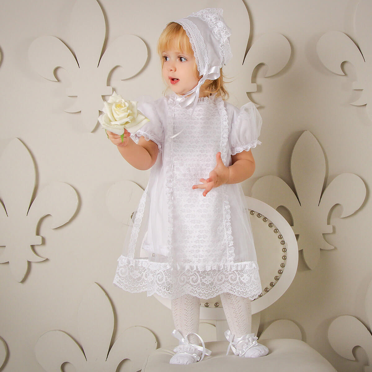 Young child in a white lace dress and Baby Baptism Bonnet holding a white flower against a decorative white background.