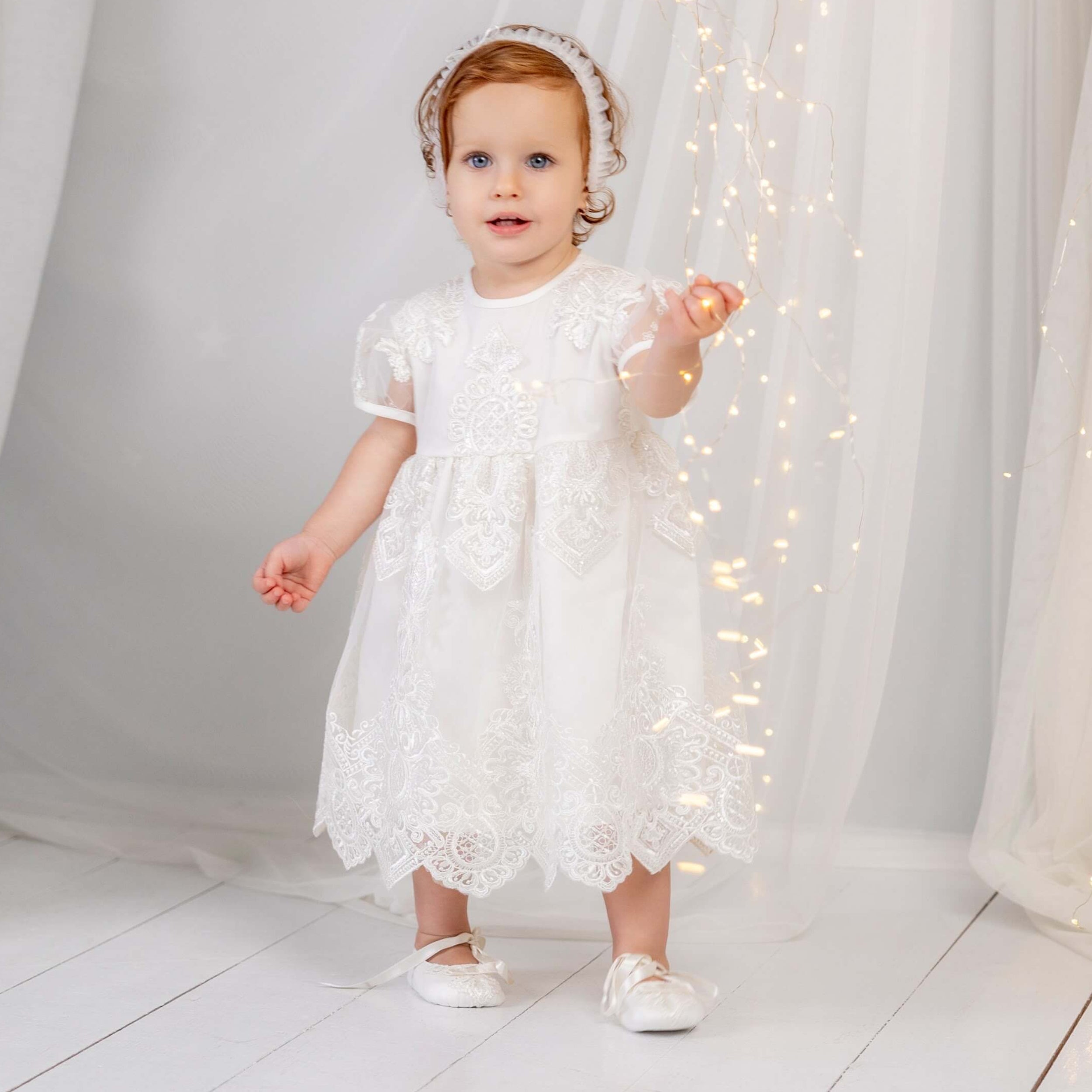 Baby in a white lace dress standing in front of white curtains with string lights.