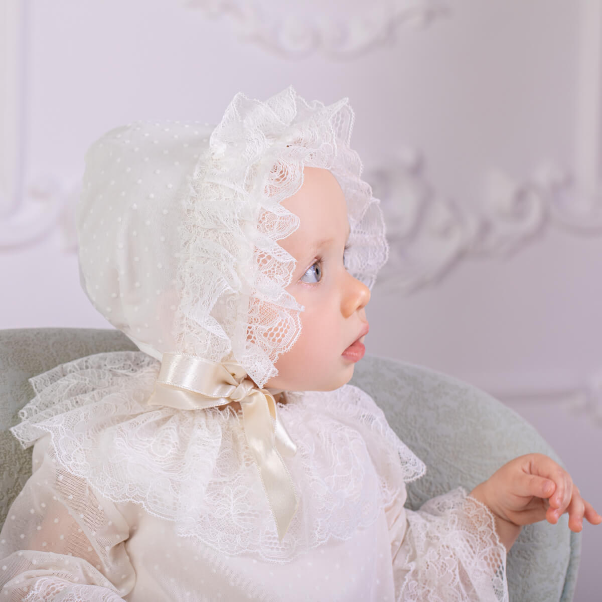 Baby wearing a white lace bonnet and  Christening Outfit against a light gray background