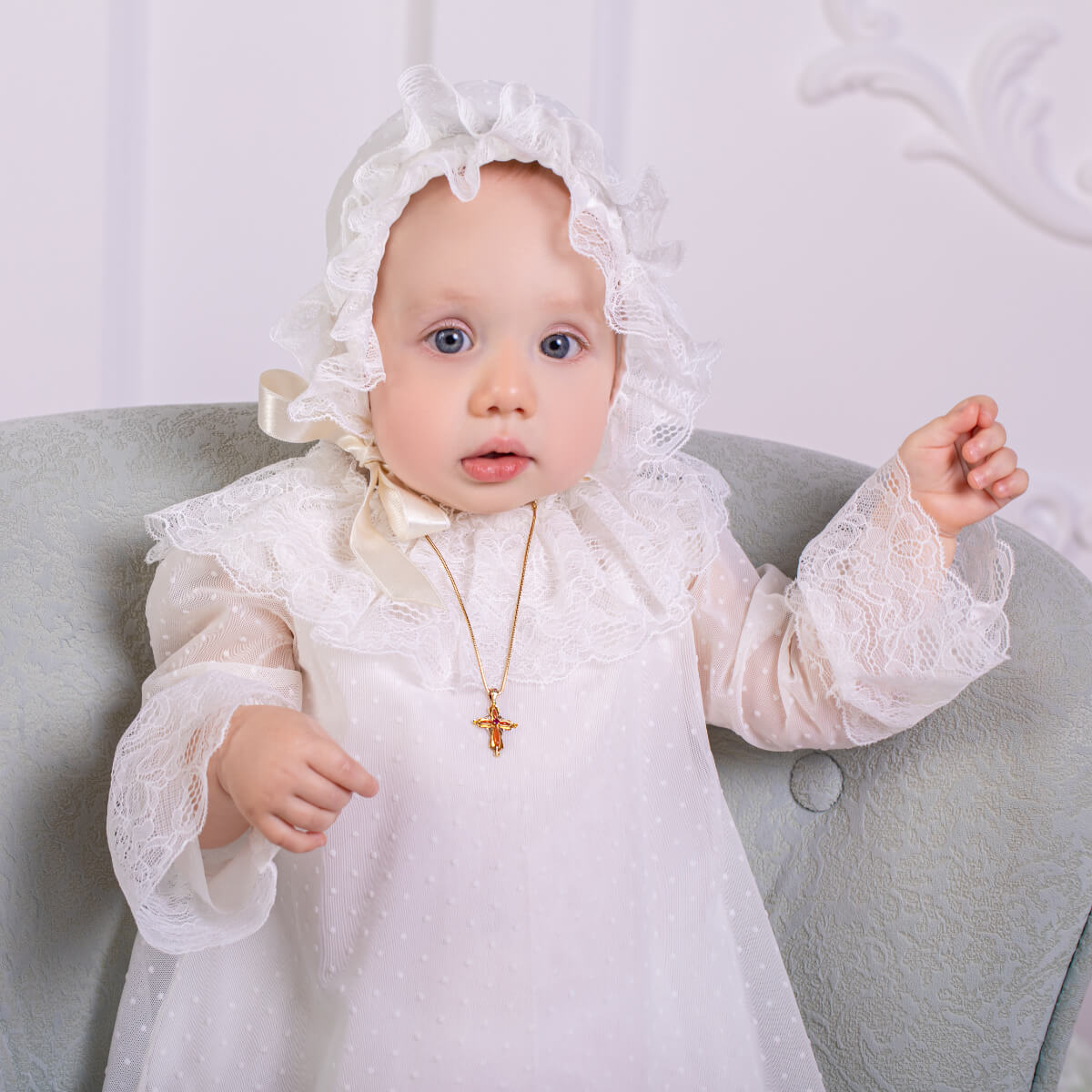 Baby in a  Christening Outfit with a bonnet sitting on a light gray couch.