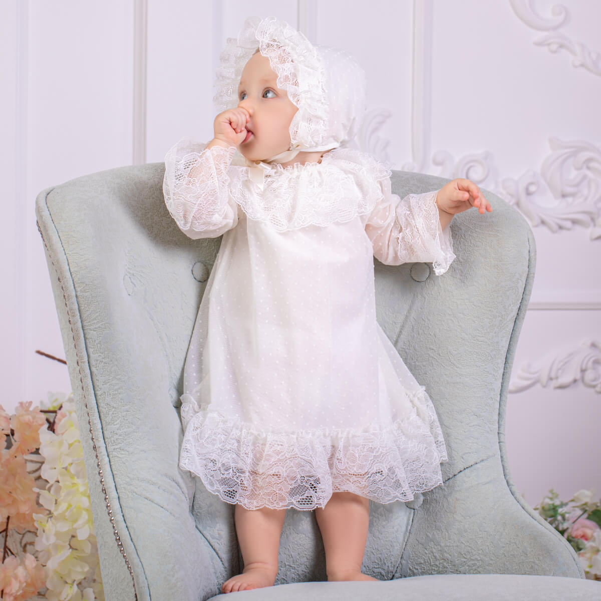 Baby in a  Christening Outfit and bonnet sitting on a gray chair with a floral background.