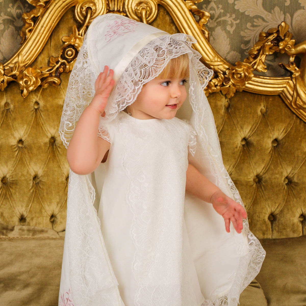 Child in a Lace baptism blanket  standing in front of an ornate gold mirror.