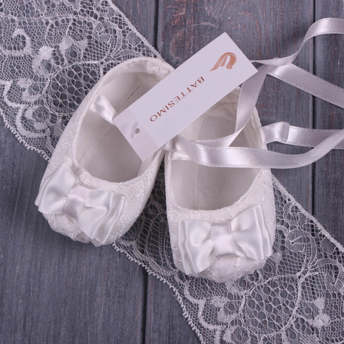 Christening Booties with a bow and ribbon on a lace surface, featuring a brand tag.