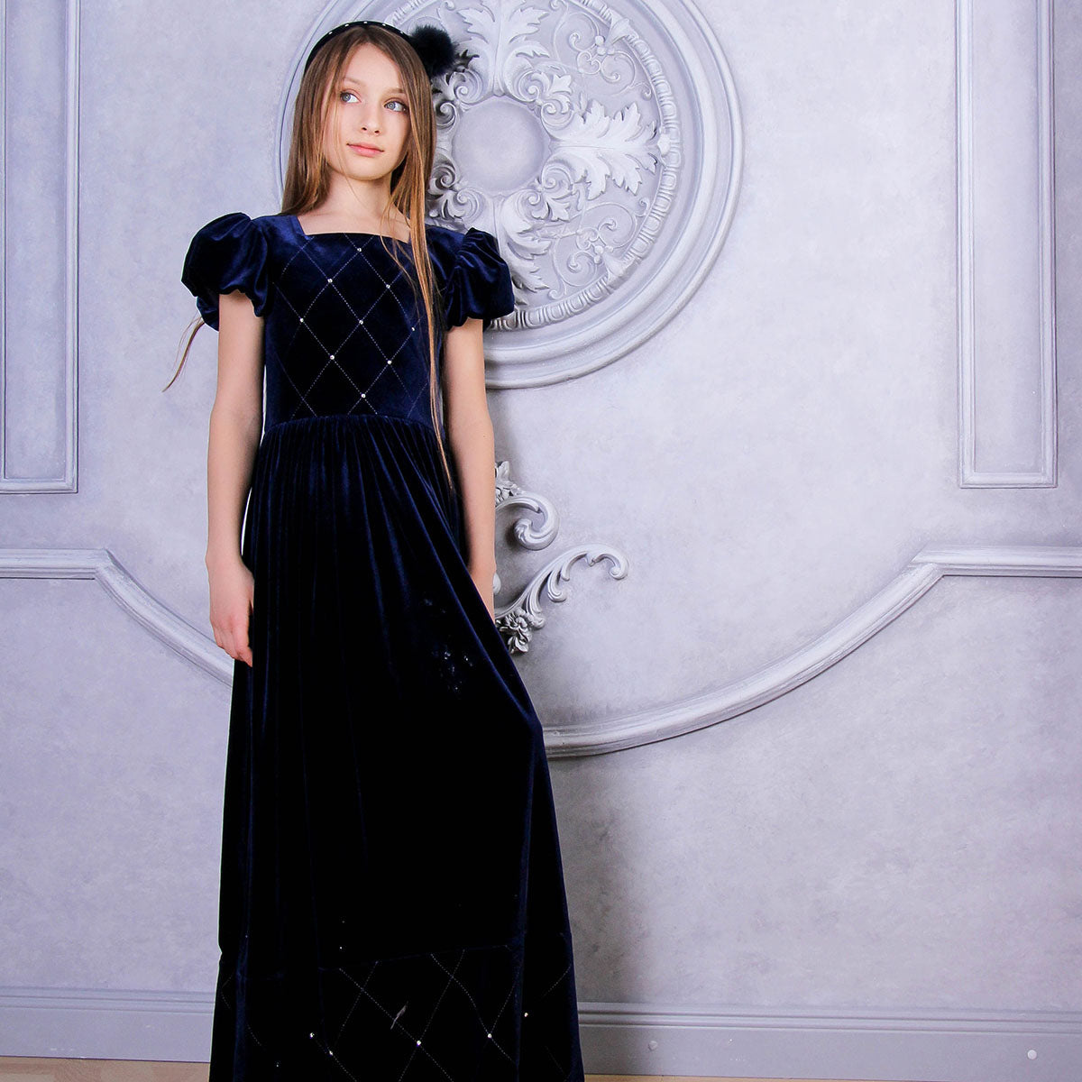 Young girl wearing a dark blue velvet dress with a decorative wall in the background