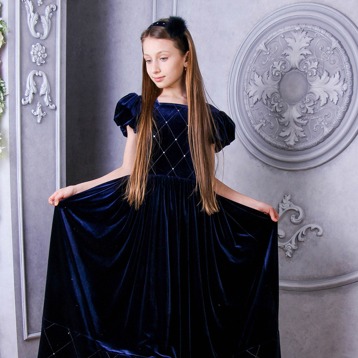 Young girl wearing a blue velvet dress against an ornate wall.