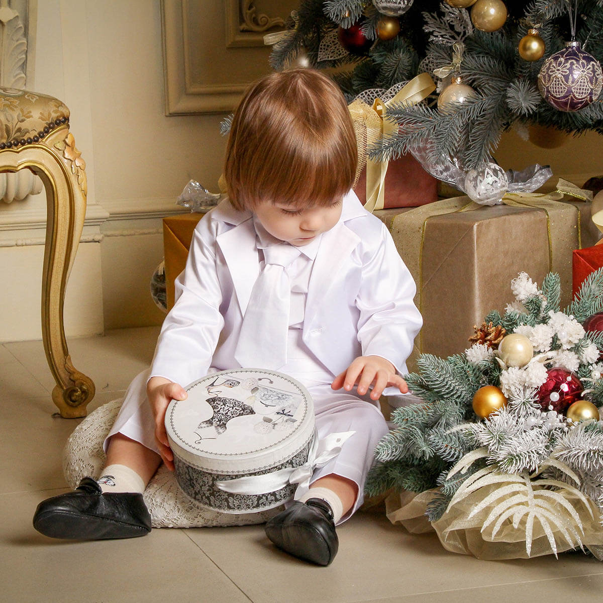Child in Baby Boy Baptism Suit  holding a gift box near a decorated Christmas tree.