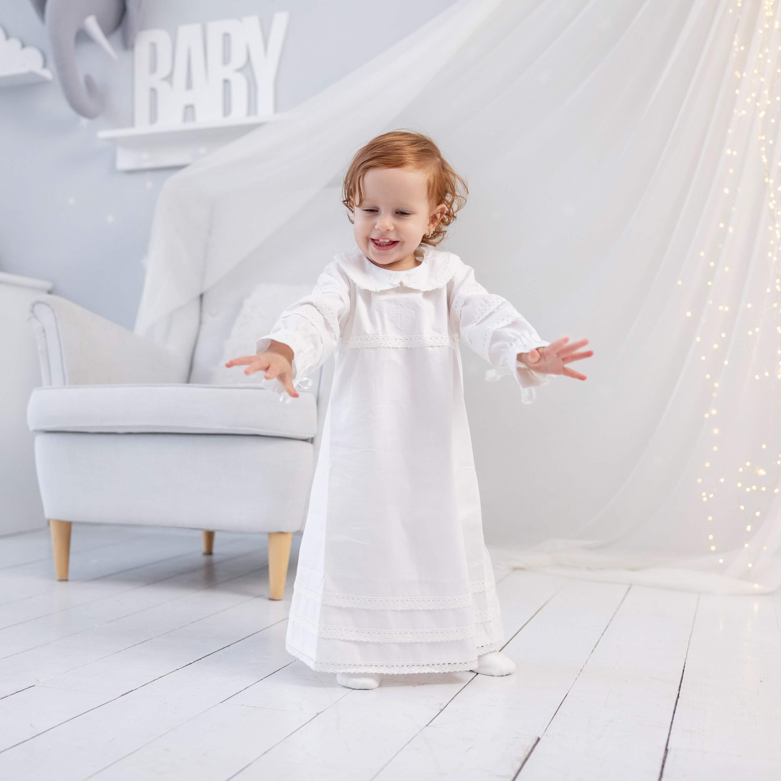 Child in a  Baptism Gown standing in a room with a white chair and decorative lights.