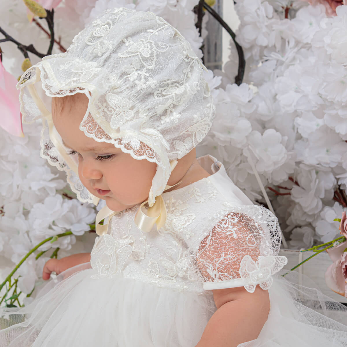 Baby in a white lace dress and vintage baby bonnet sitting among white flowers