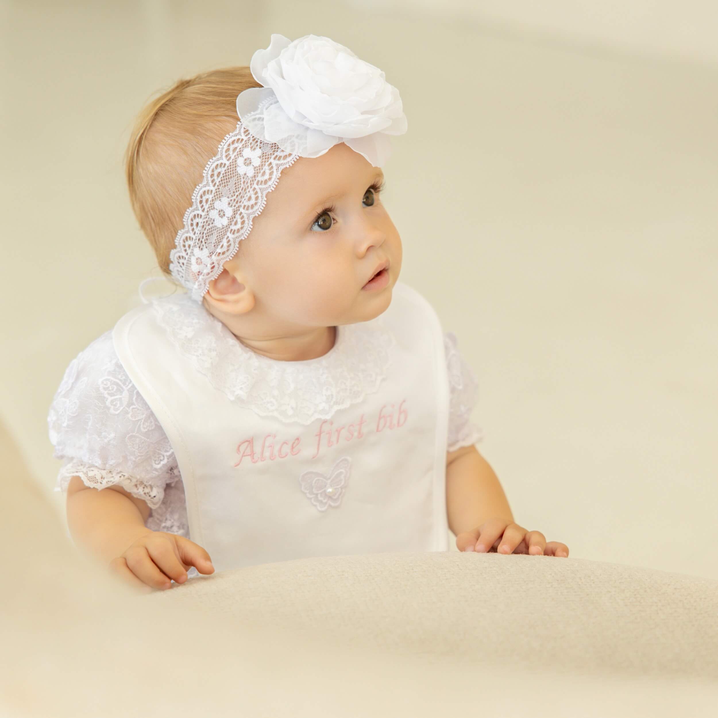 Baby wearing a Baptism Bib and headband with a flower, sitting on a beige surface.