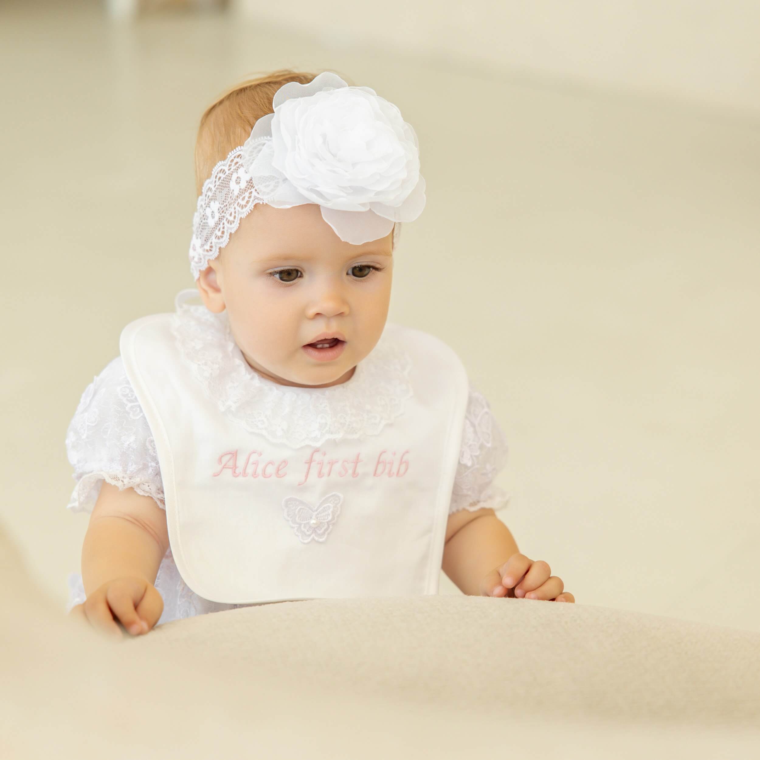 Baby wearing a Baptism Bib with 'Alice first bib' text and a matching headband on a beige background