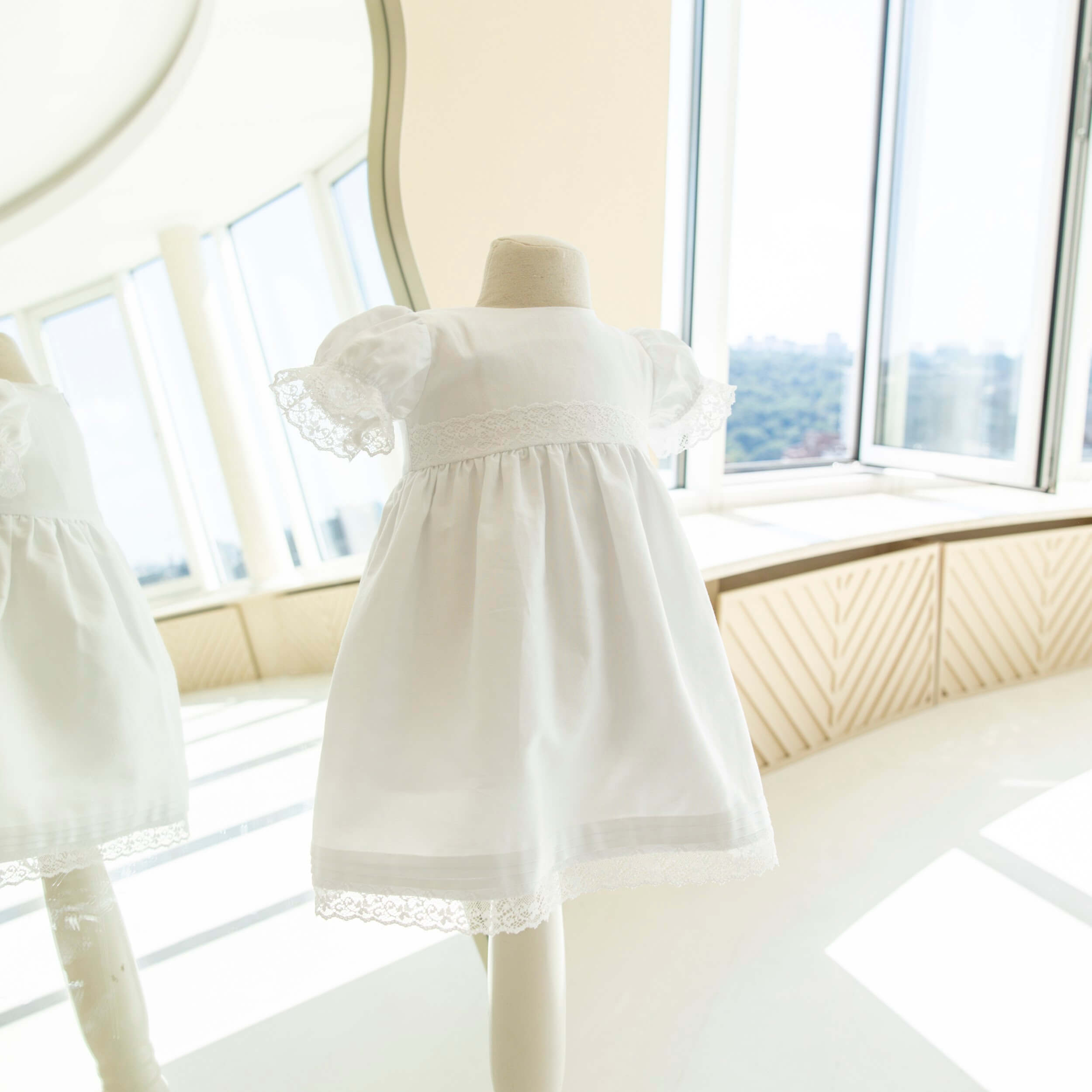 Baptism Dress with Lace on a mannequin in a bright room with large windows.