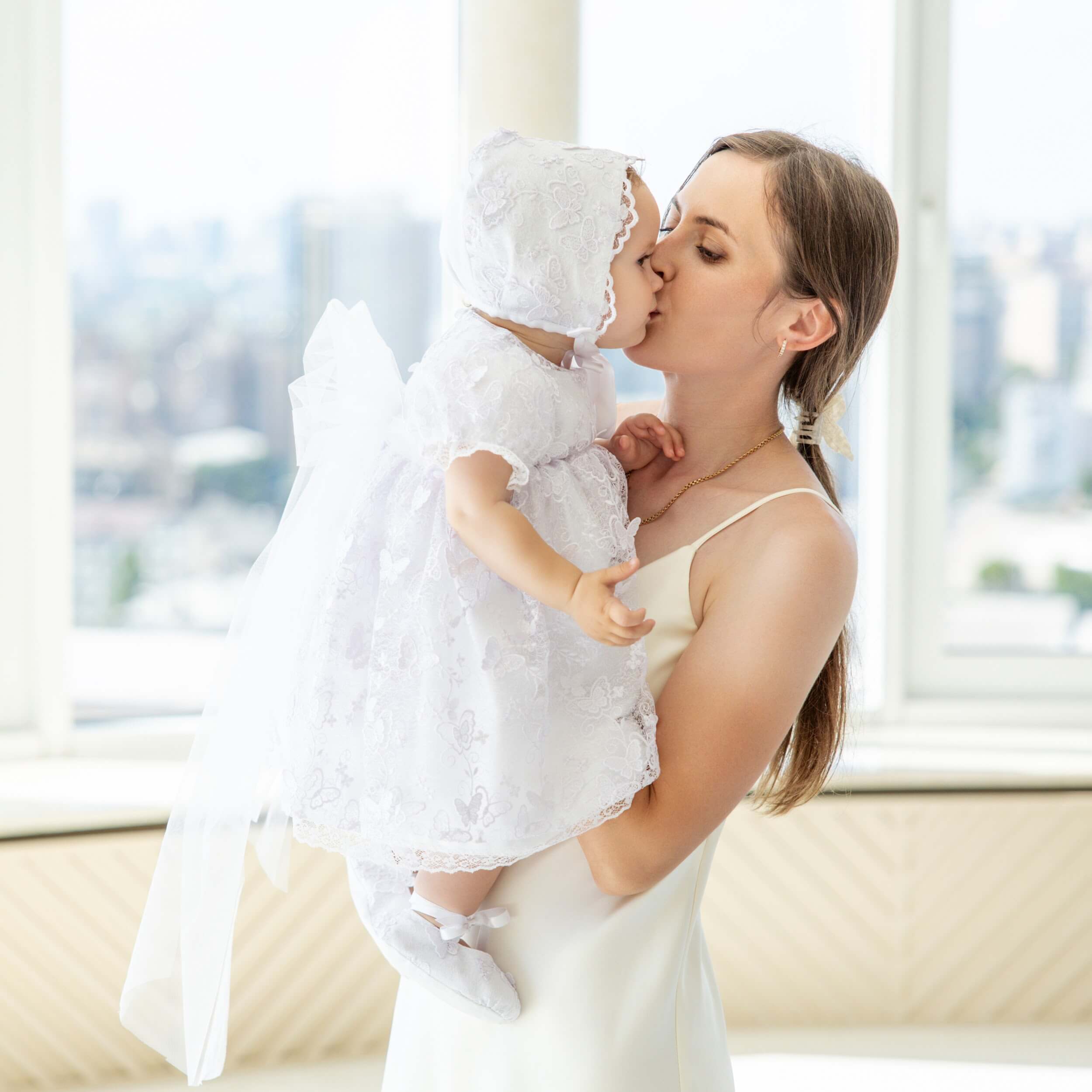 Woman holding a baby in a Baptism Dress with Lace by a window with a cityscape view.