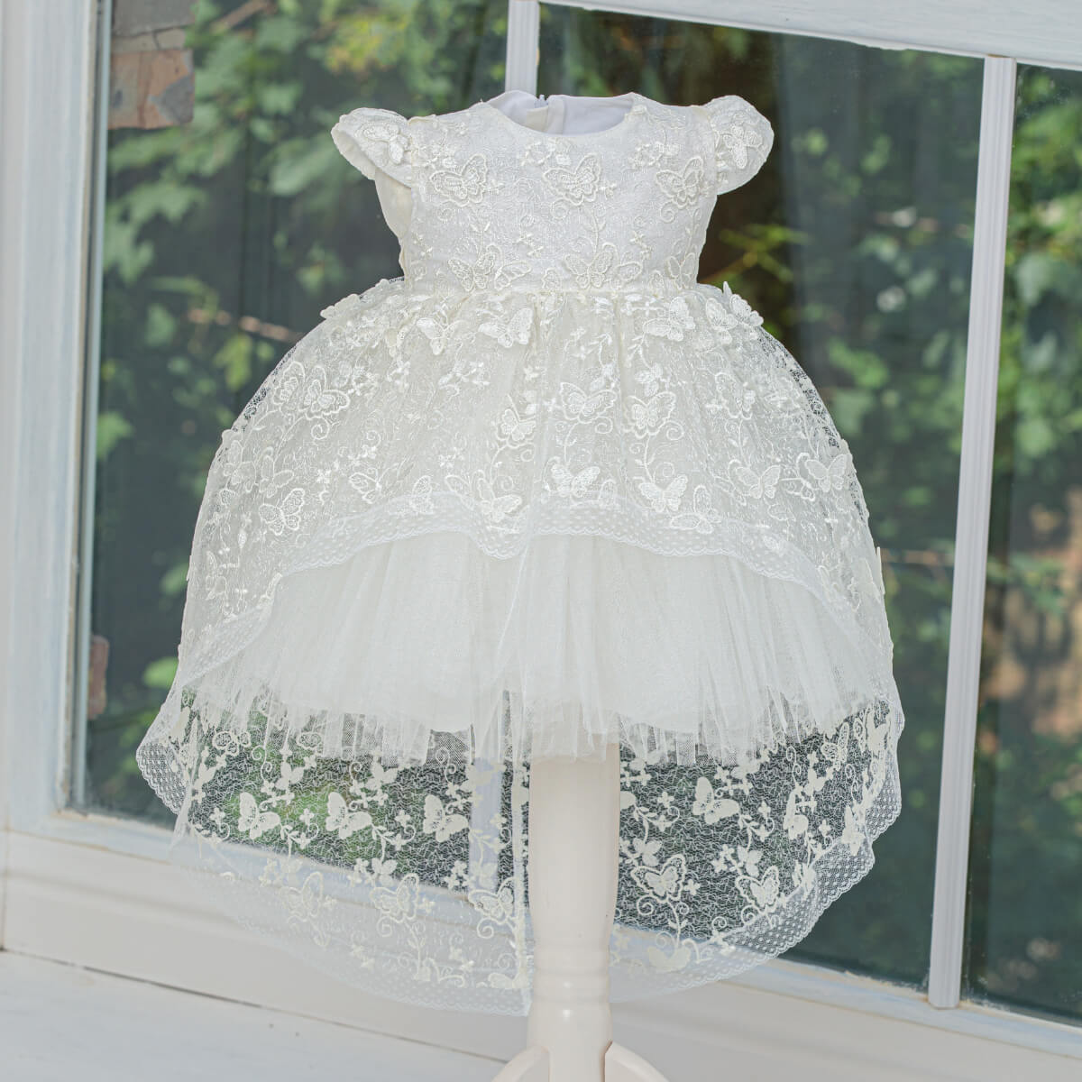 Baptism Dress with Lace on a mannequin in front of a window with greenery outside