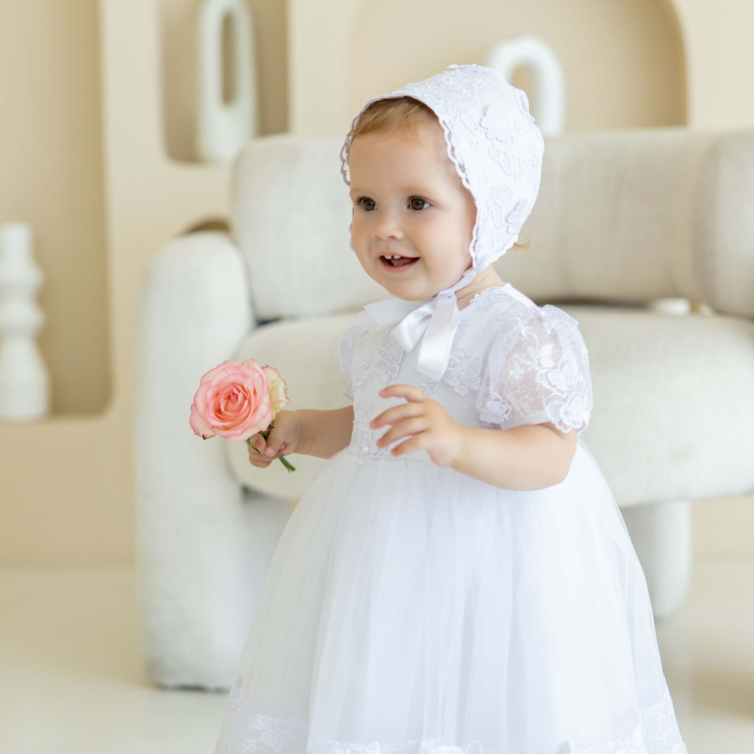 Baptism Dress with Butterfly Lace and bonnet holding a pink flower, sitting on a beige couch.