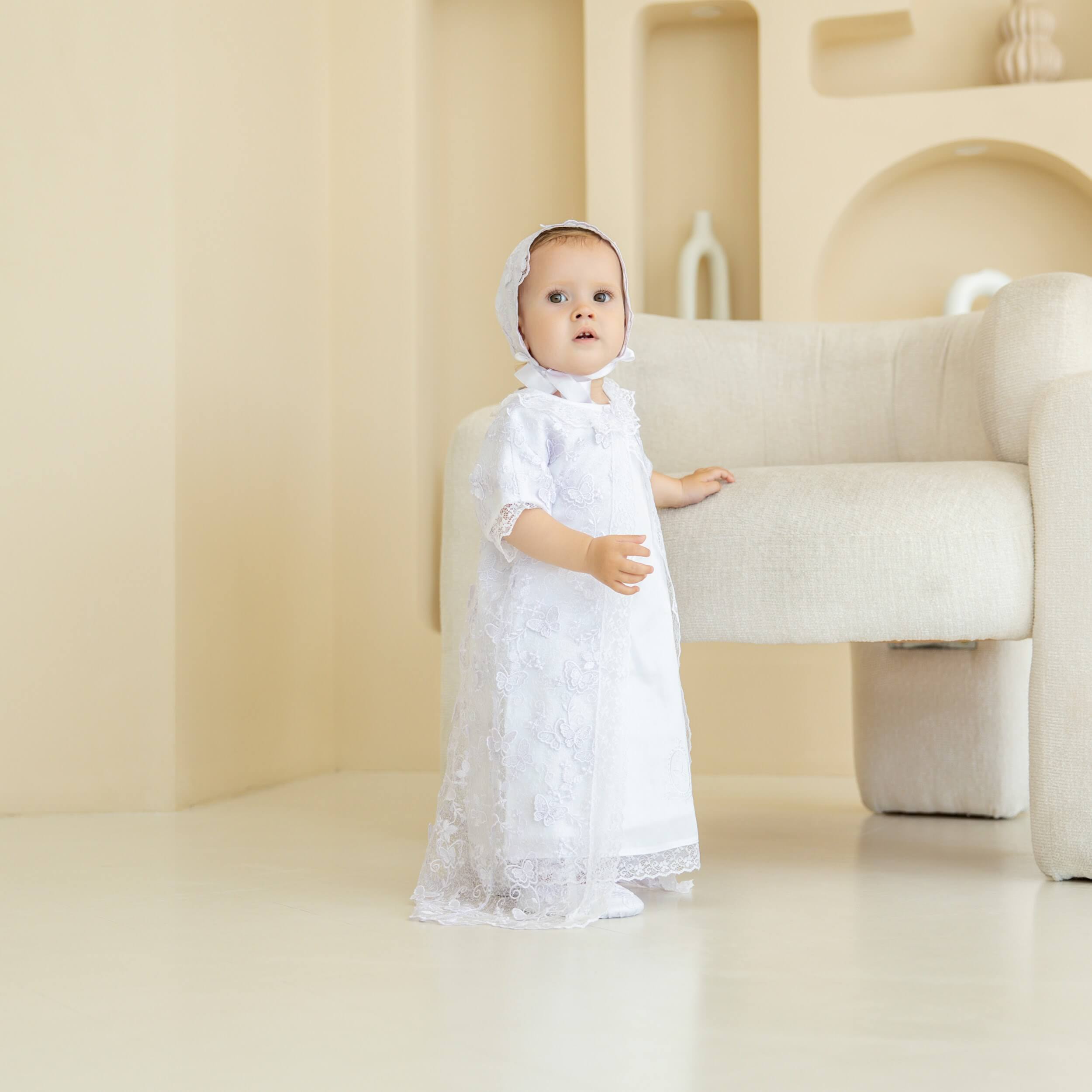 Baby in a  Christening Gown for Girls standing in a minimalistic room with a beige sofa.