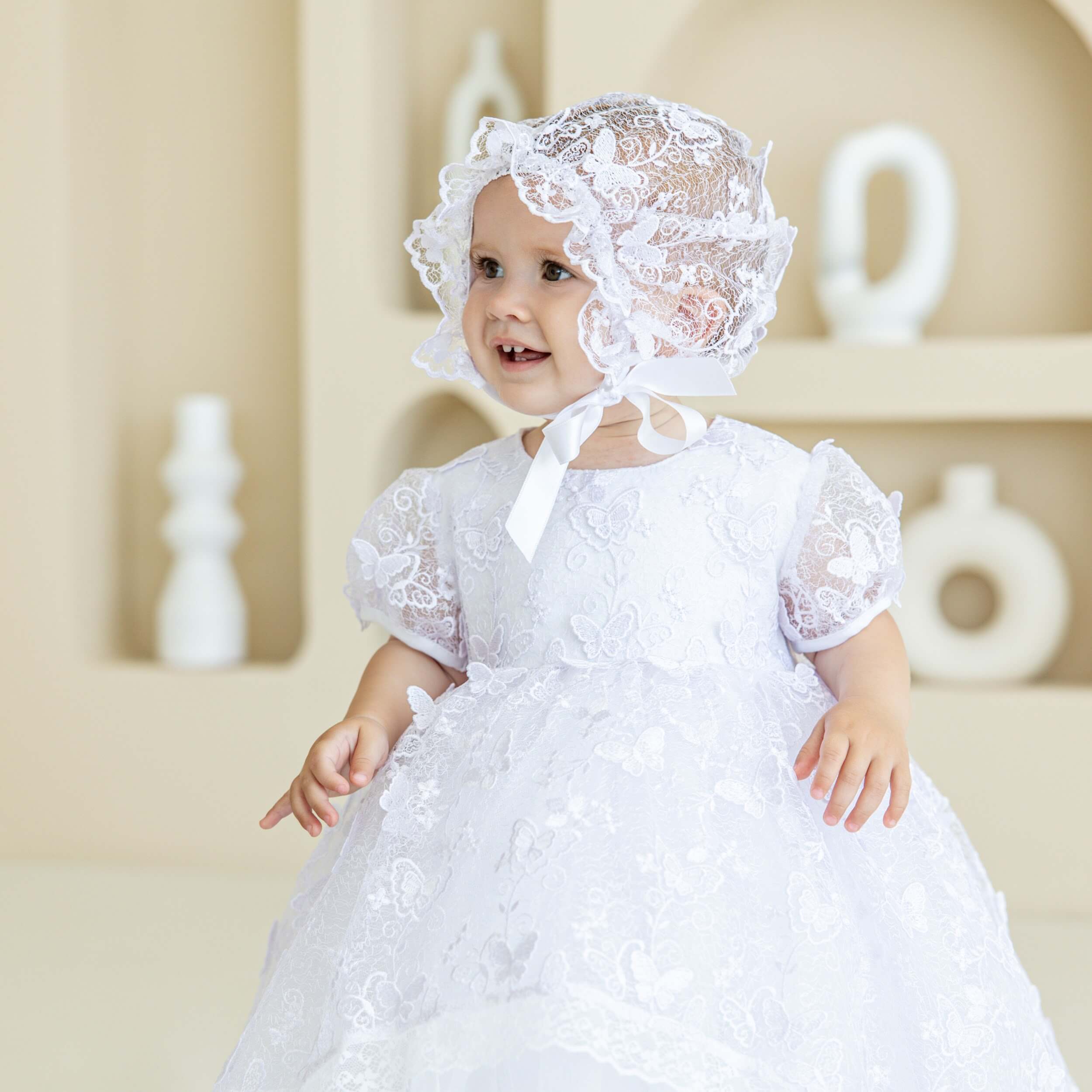 Baby in a white lace dress and vintage baptism bonnet standing against a neutral background