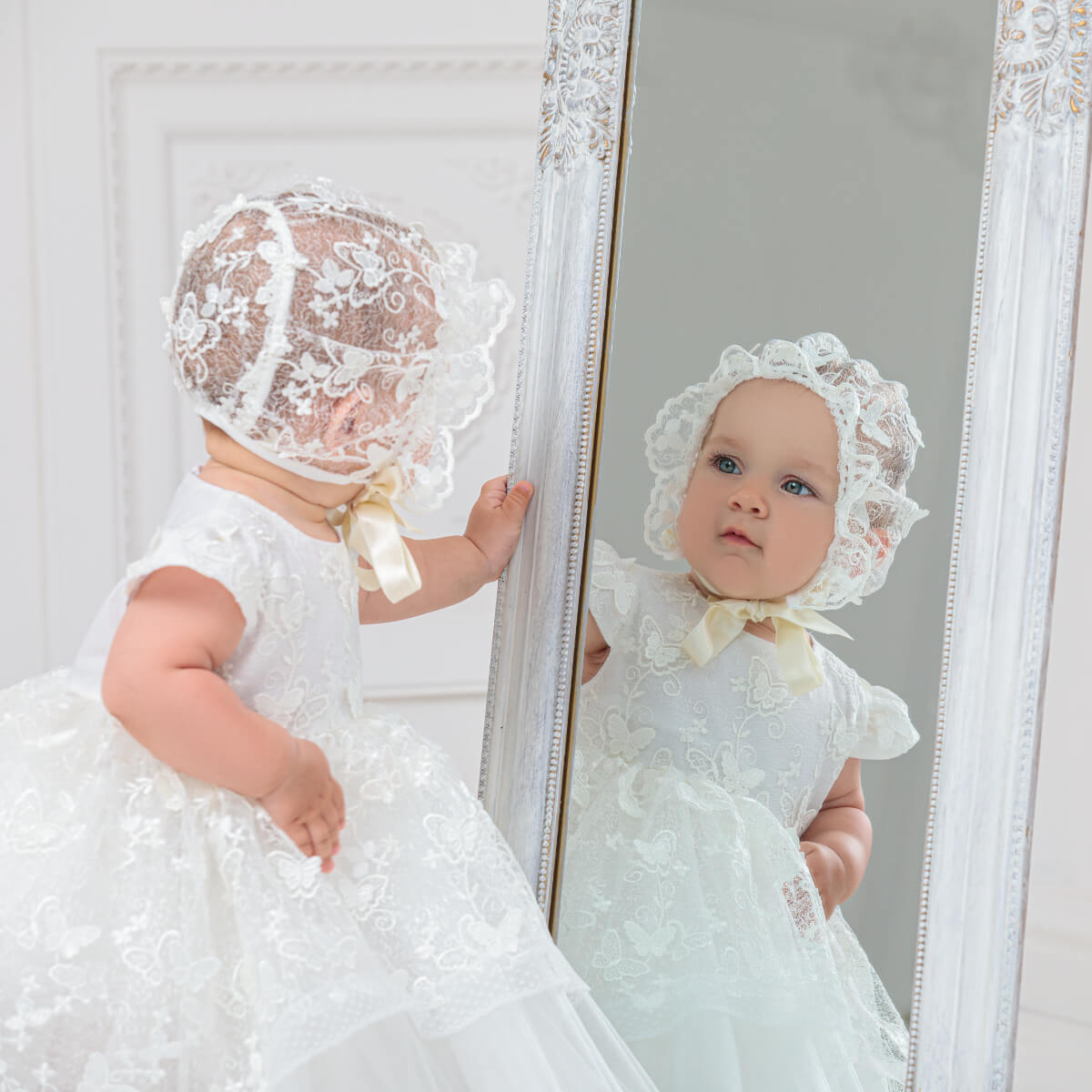 Baby in a white lace dress and Lace Baptism Bonnet standing in front of a mirror.