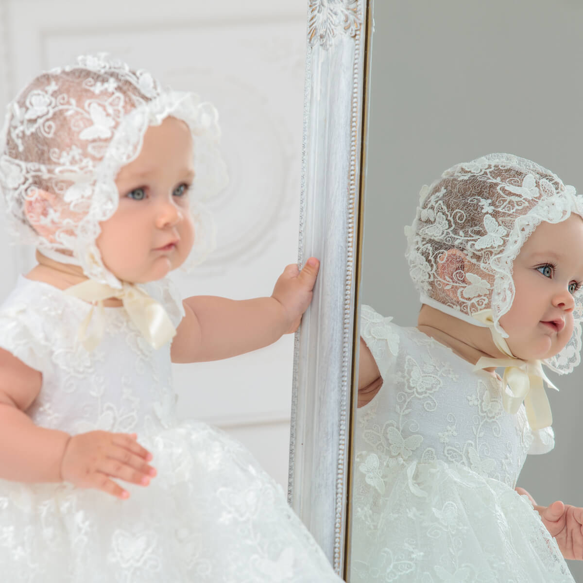 Baby in a Lace Baptism Bonnet and dress standing in front of a mirror.