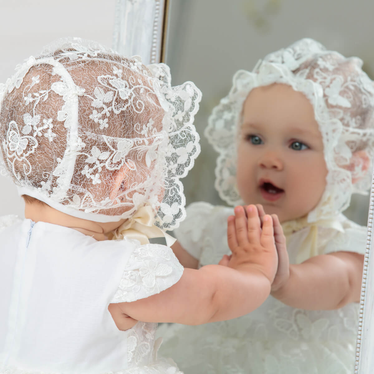 Baby wearing a Lace Baptism Bonnet looking at its reflection in a mirror.