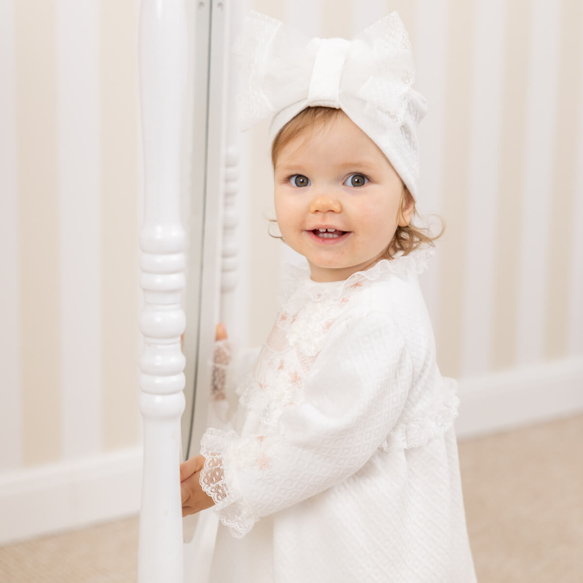 Baby in a white outfit with a Lace Christening Bonnet standing in front of a white curtain.