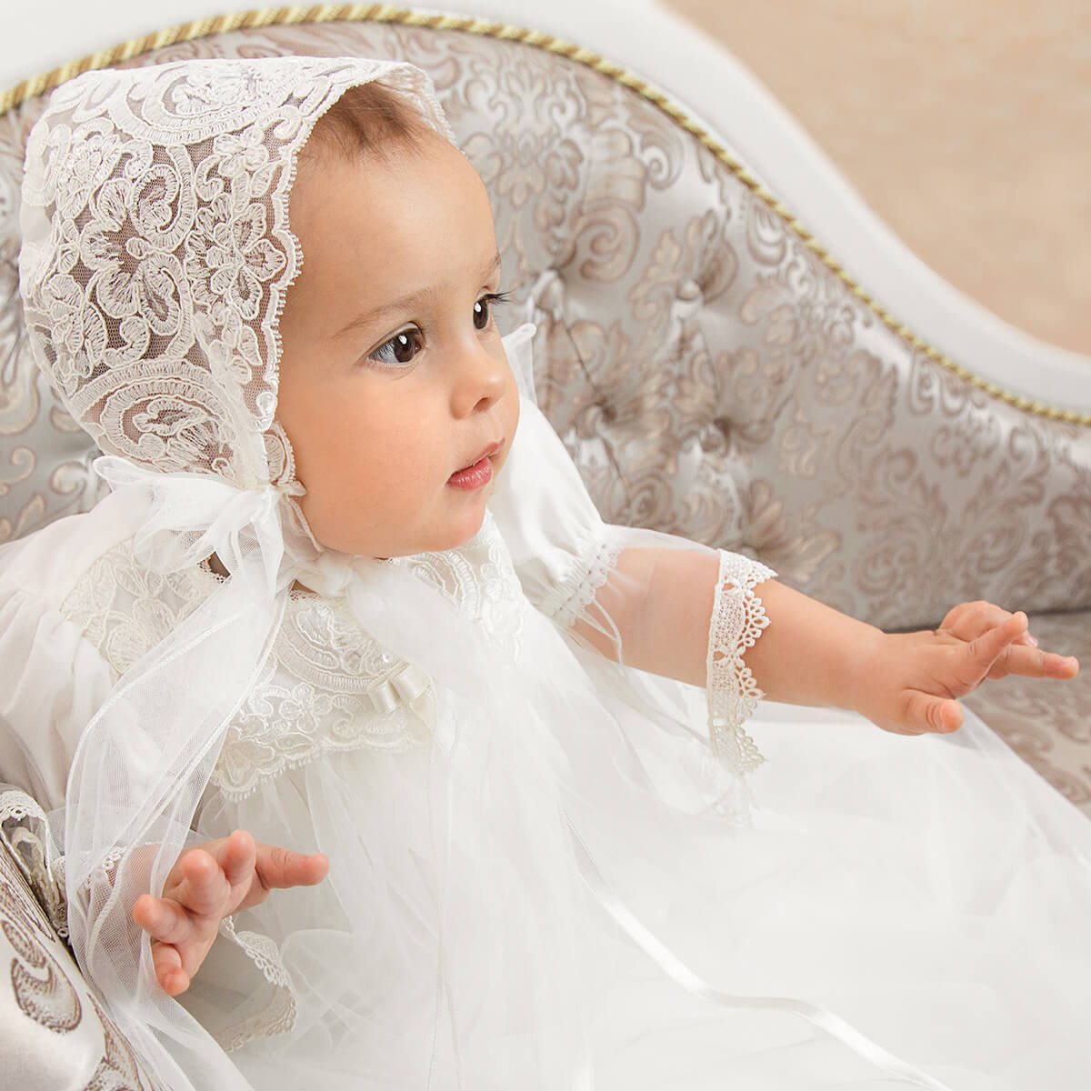 Baby in a Christening Gown with Train for Girls sitting on a decorative chair.